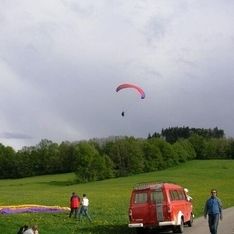 Ein Paragleiter fliegt über einer Wiese mit roten Auto im Vordergrund