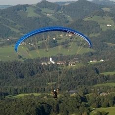 Ein Paragleiter mit blauem Schirm im Flug vor Wald im Hintergrund